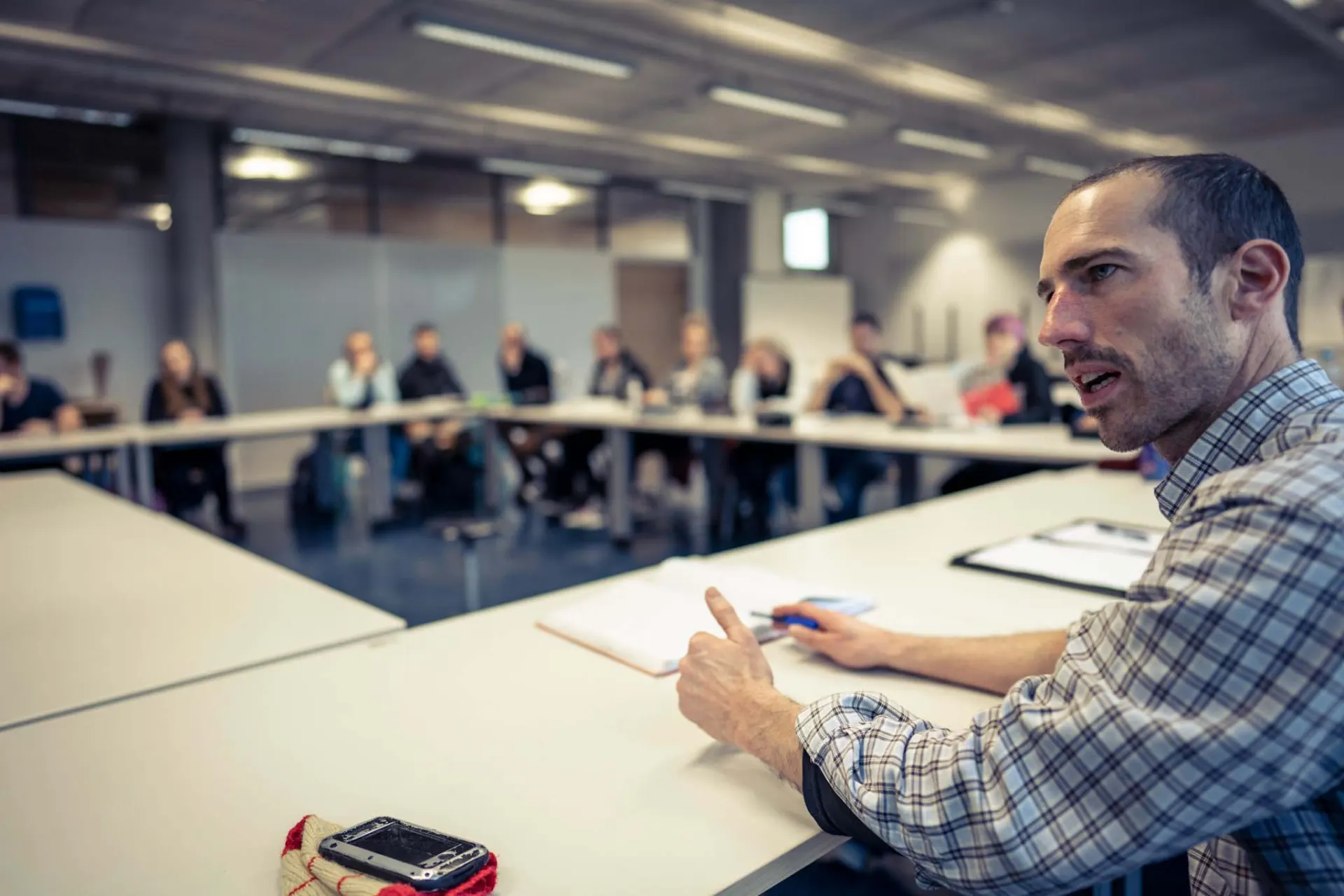 A workshop: a facilitator speaks while a group listens around tables in the background.